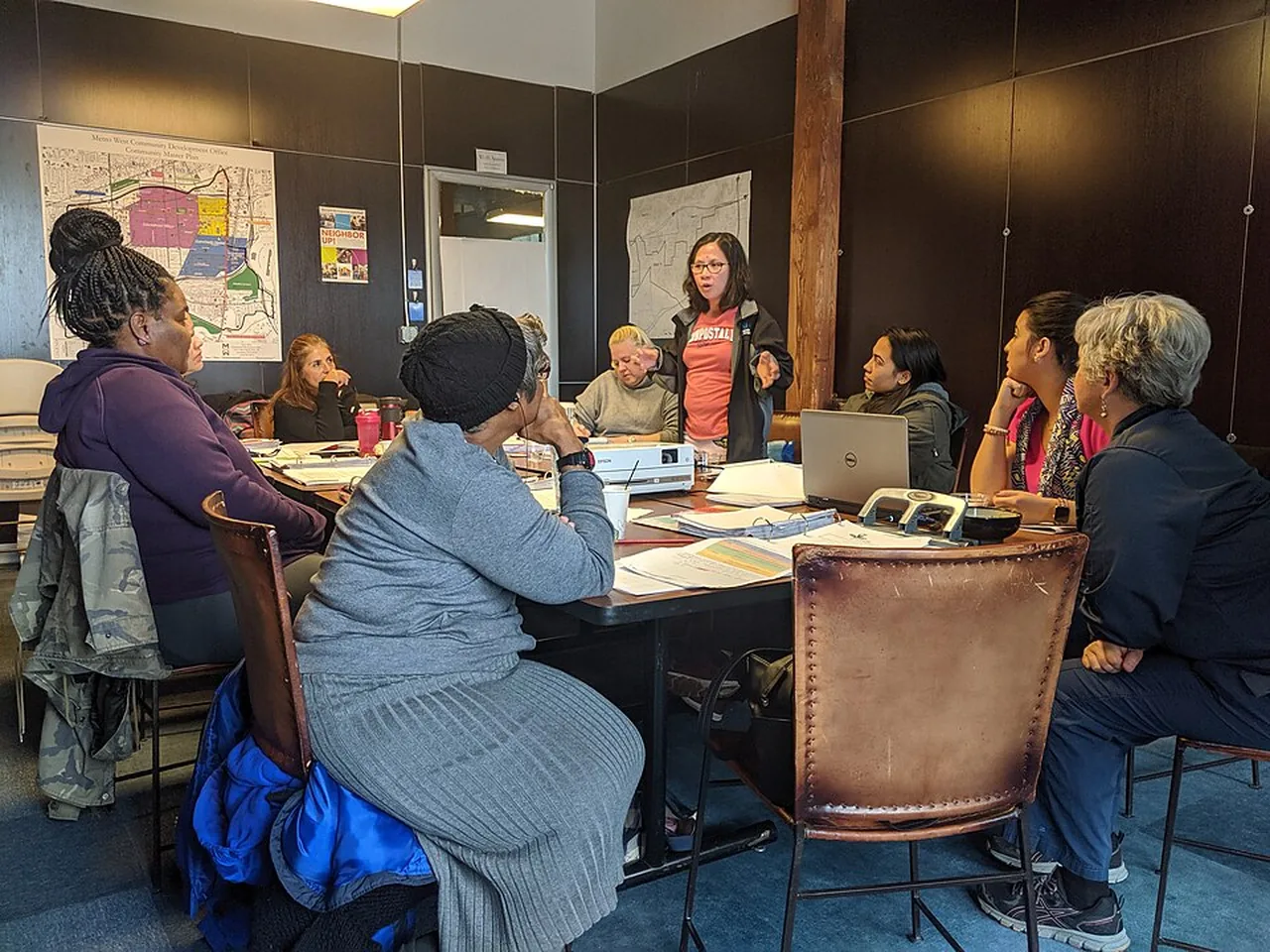 A group of women sits around a table while one participant stands and speaks.